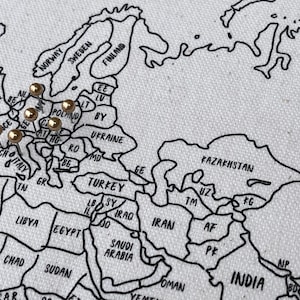 May include: A white fabric map featuring black outlines of countries and continents with text labels. Several gold-colored push pins are inserted into the European region of the map, marking specific locations across various countries.