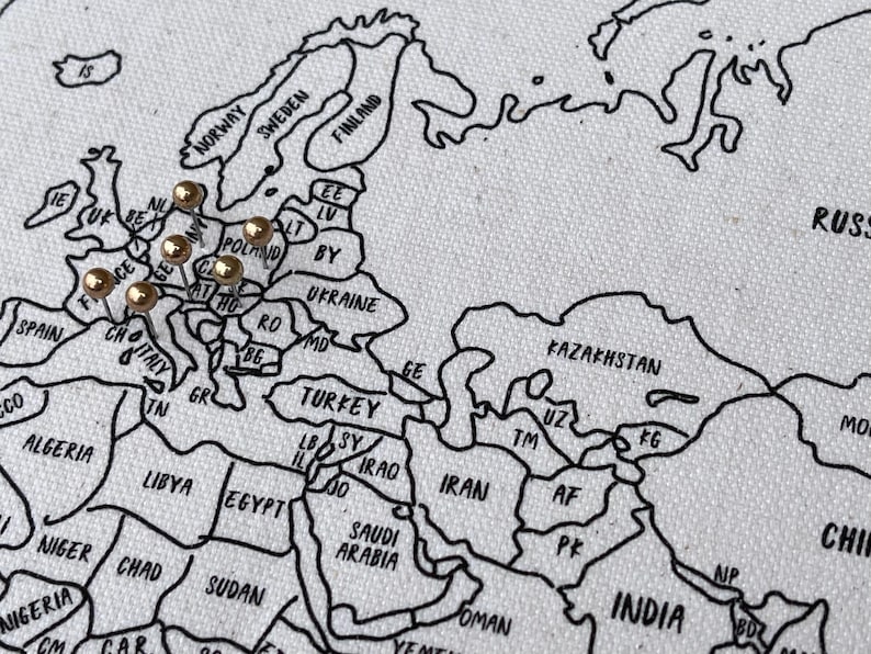 May include: A white fabric map featuring black outlines of countries and continents with text labels. Several gold-colored push pins are inserted into the European region of the map, marking specific locations across various countries.
