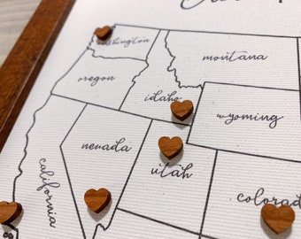 May include: A framed canvas map of the western United States, with state outlines and handwritten state names. Small, heart-shaped wooden pins mark locations on the map. The frame is made of dark brown wood.