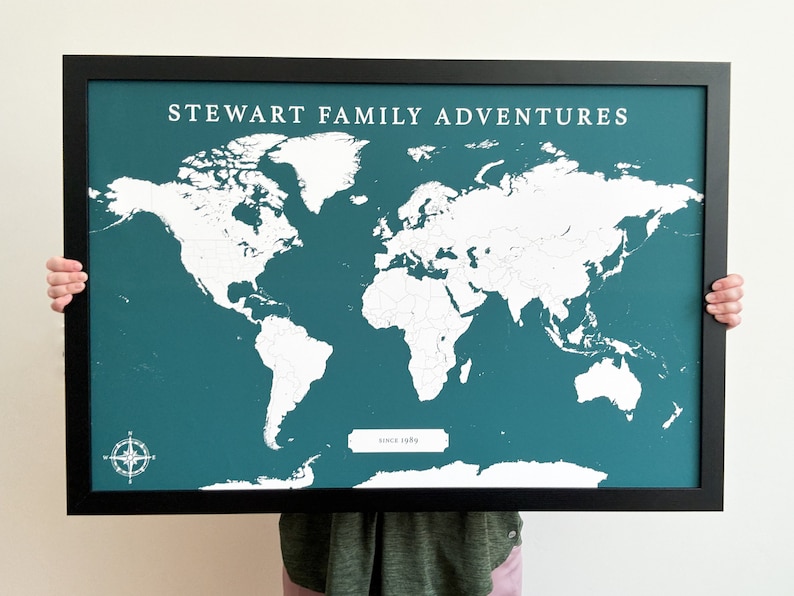 May include: A teal blue world map in a black frame with the text "STEWART FAMILY ADVENTURES since 1989" printed on the map. The map is blank and ready to be marked with pins.