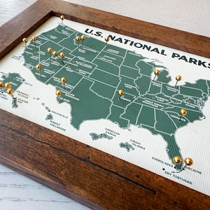 Pu&ograve; includere: Una mappa incorniciata dei parchi nazionali statunitensi con puntine dorate che segnano le localit&agrave;. La mappa &egrave; verde con testo bianco ed &egrave; inserita in una cornice di legno marrone scuro. Il testo "U.S. NATIONAL PARKS" &egrave; in alto.