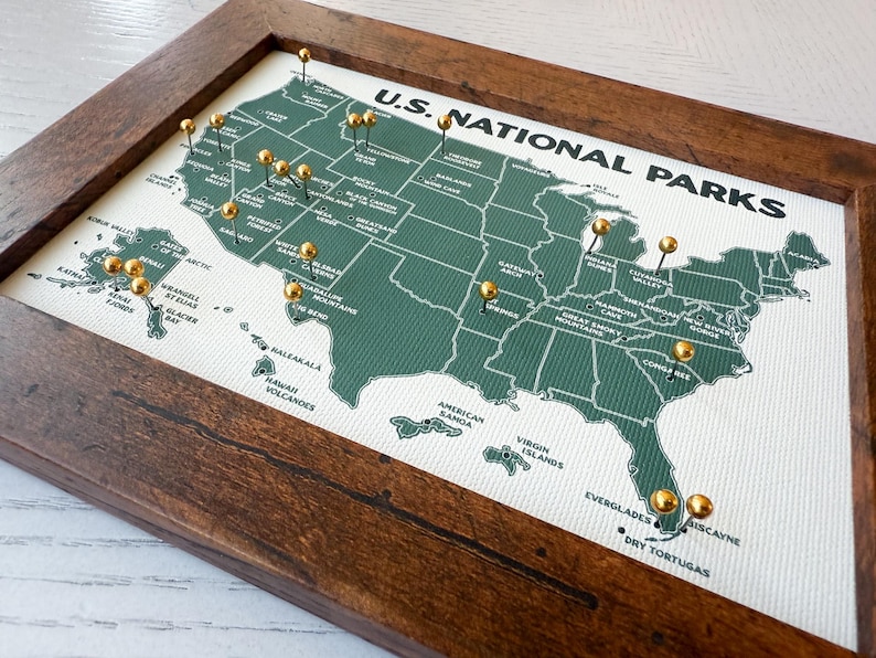 Pu&ograve; includere: Una mappa incorniciata dei parchi nazionali statunitensi con puntine dorate che segnano le localit&agrave;. La mappa &egrave; verde con testo bianco ed &egrave; inserita in una cornice di legno marrone scuro. Il testo "U.S. NATIONAL PARKS" &egrave; in alto.
