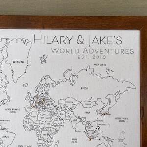 May include: A framed world map on a light-colored fabric background with black outlines of continents and countries. The text reads HILARY & JAKE'S WORLD ADVENTURES EST. 2010. Small metallic pins are inserted into various locations on the map.