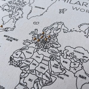 May include: A white canvas world map with black printed outlines of continents and countries. The map features gold-colored push pins inserted into various locations across Europe and North America. The text HILARY & WORLD EST is visible in the top right corner.