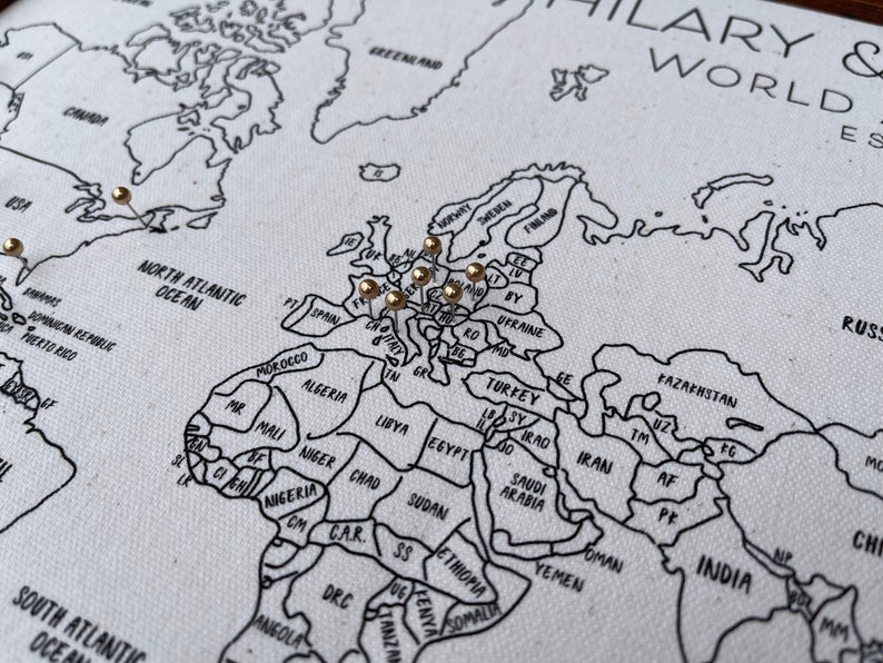 May include: A white canvas world map with black printed outlines of continents and countries. The map features gold-colored push pins inserted into various locations across Europe and North America. The text HILARY & WORLD EST is visible in the top right corner.