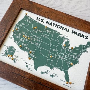 Pu&ograve; includere: Una mappa incorniciata dei parchi nazionali degli Stati Uniti. La mappa &egrave; verde con testo bianco e puntine dorate che segnano le posizioni dei parchi. La cornice &egrave; in legno marrone scuro. Le parole "U.S. NATIONAL PARKS" sono stampate in alto.