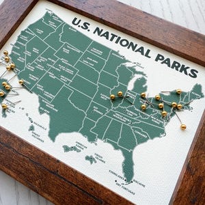 Pu&ograve; includere: Una mappa incorniciata dei parchi nazionali degli Stati Uniti. La mappa &egrave; verde con testo bianco e puntine dorate che segnano le posizioni. La cornice &egrave; in legno marrone scuro. Le parole "U.S. NATIONAL PARKS" sono stampate in alto.