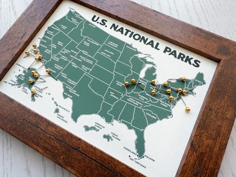Pu&ograve; includere: Una mappa incorniciata dei parchi nazionali degli Stati Uniti. La mappa &egrave; verde con testo bianco e puntine dorate che segnano le posizioni. La cornice &egrave; in legno marrone scuro. Le parole "U.S. NATIONAL PARKS" sono stampate in alto.
