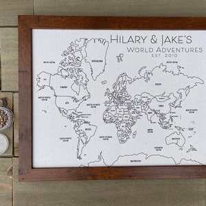 May include: A framed world map print on white fabric with black outlines and text reading HILARY & JAKE'S WORLD ADVENTURES EST. 2010. The map features labeled continents and oceans, accompanied by a small clear container filled with metallic push pins.