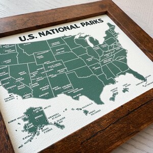 Pu&ograve; includere: Una mappa incorniciata dei parchi nazionali degli Stati Uniti. La mappa &egrave; verde su sfondo bianco, con ogni parco etichettato. La cornice &egrave; in legno marrone scuro. Le parole "U.S. NATIONAL PARKS" sono stampate in alto.