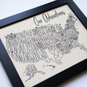 May include: A framed map of the United States with the states spelled out in a stylized font. The map is decorated with colorful push pins, and the title "Our Adventures" is written at the top.