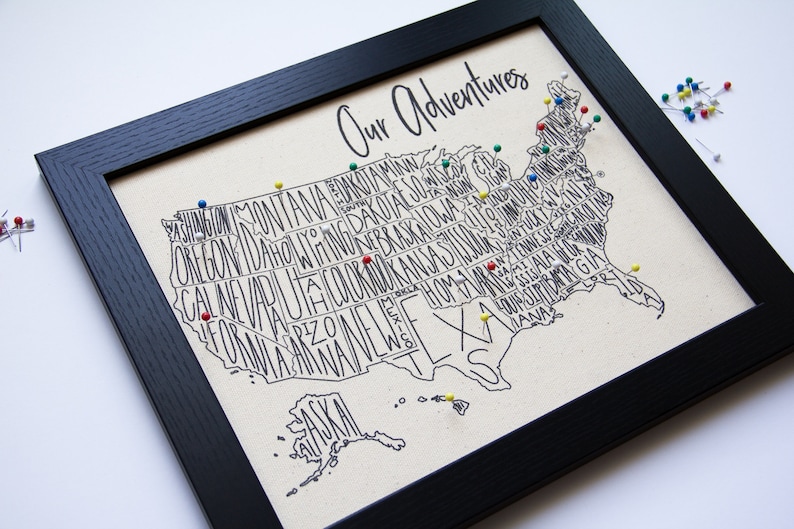 May include: A framed map of the United States with the states spelled out in a stylized font. The map is decorated with colorful push pins, and the title "Our Adventures" is written at the top.