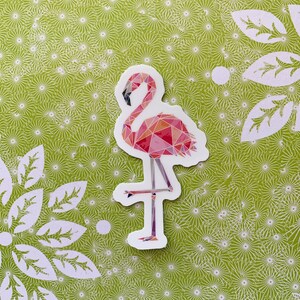 Puede incluir: Una pegatina de flamenco rosa con un dise&ntilde;o geom&eacute;trico. El flamenco est&aacute; de pie sobre una pierna con la cabeza inclinada hacia abajo. La pegatina est&aacute; sobre un fondo verde con un patr&oacute;n floral blanco.