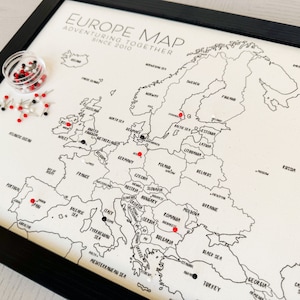 May include: A black and white map of Europe with red and white push pins marking locations. The map is framed in black and has the text "Europe Map Adventuring Together Since 2010" at the top.