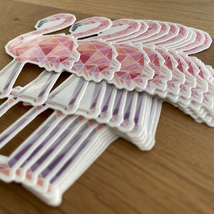 Puede incluir: Una pila de pegatinas de flamencos rosas con un dise&ntilde;o geom&eacute;trico. Las pegatinas est&aacute;n hechas de un material brillante y tienen un fondo blanco.