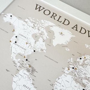 May include: A framed world map with the words "WORLD ADVENTURE" in dark brown. The map is light beige with black and gold push pins marking locations. The frame is white.
