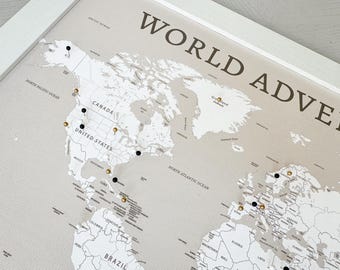 May include: A framed world map with the words "WORLD ADVENTURE" in dark brown. The map is light beige with black and gold push pins marking locations. The frame is white.