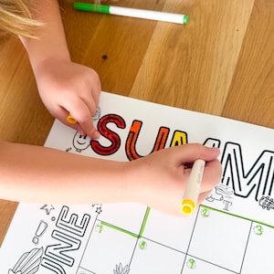 May include: A child's hands coloring the word "SUMMER" on a white paper calendar with black outlines. The word "SUMMER" is in orange and red. A green and white marker is on the wooden surface.