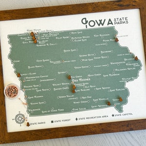May include: A green and white map of Iowa with wooden markers placed on each state park. The map is framed in a dark brown wood frame. The text "Iowa State Parks" is at the top of the map.
