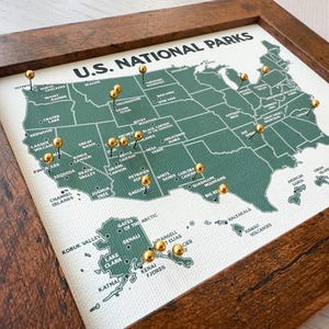 Pu&ograve; includere: Una mappa incorniciata dei parchi nazionali degli Stati Uniti con un contorno verde degli Stati Uniti. Spilli dorati segnano varie localit&agrave; dei parchi. La mappa &egrave; etichettata "U.S. NATIONAL PARKS" in testo bianco ed &egrave; incorniciata in una cornice di legno marrone scuro.
