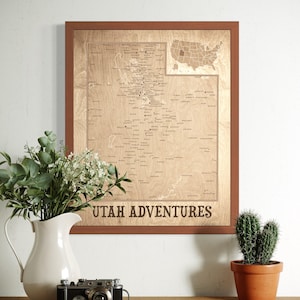 May include: A framed wooden map of Utah with the text "UTAH ADVENTURES" at the bottom. The map is light brown with dark brown text and a dark brown frame. A small inset map of the United States is in the upper right corner.