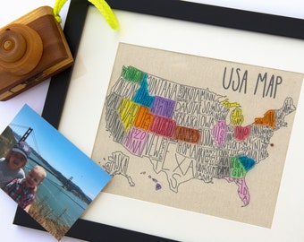 May include: A framed map of the United States with each state outlined and labeled with its name in colorful handwritten text. The map is on a beige canvas background and the frame is black.