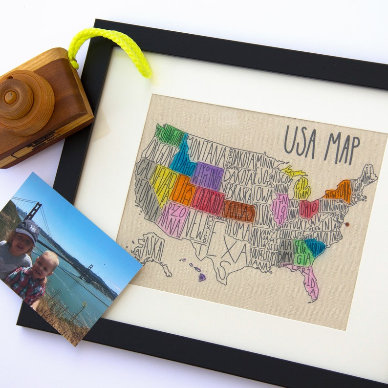 May include: A framed map of the United States with each state outlined and labeled with its name in colorful handwritten text. The map is on a beige canvas background and the frame is black.