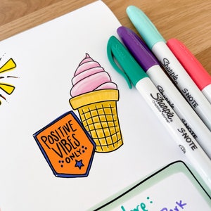 May include: A white sheet of paper with a hand-drawn ice cream cone, a sun, and a sticker that says "Positive Vibes Only". There are four colorful Sharpie S-Note markers on the paper.