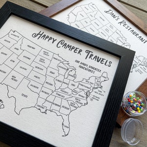 Op de afbeelding: Twee ingelijste kaarten van de Verenigde Staten met de tekst "Happy Camper Travels Our Daddy, Daughter Adventures" en "Dan's Restaurant Tour" erop gedrukt. De kaarten zijn wit met zwarte contouren van de staten. Er zijn duimspijkers in een kleine container naast de kaarten.