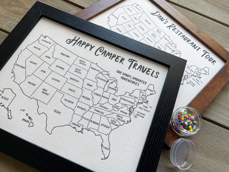 Op de afbeelding: Twee ingelijste kaarten van de Verenigde Staten met de tekst "Happy Camper Travels Our Daddy, Daughter Adventures" en "Dan's Restaurant Tour" erop gedrukt. De kaarten zijn wit met zwarte contouren van de staten. Er zijn duimspijkers in een kleine container naast de kaarten.