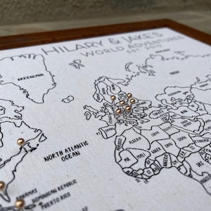 May include: A framed world map on white fabric with black outlines and text reading HILARY & JAKE'S WORLD ADVENTURES EST. 2015. The map features metallic gold push pins inserted into various locations across the continents of North America and Europe.