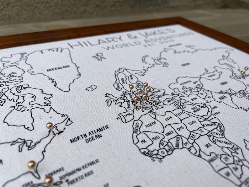 May include: A framed world map on white fabric with black outlines and text reading HILARY & JAKE'S WORLD ADVENTURES EST. 2015. The map features metallic gold push pins inserted into various locations across the continents of North America and Europe.
