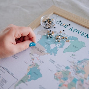 May include: A framed world map with the text "OUR ADVENTURES" in teal. A small container of map pins sits near the top, with a hand placing a pin on the United States. The map is in a light wooden frame.