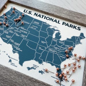 May include: A framed map of the U.S. National Parks with a dark blue map and white text. The map is covered in copper-colored push pins. The frame is a light gray color. The text "U.S. NATIONAL PARKS" is at the top of the map.