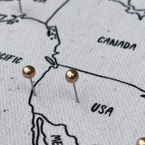 May include: A white fabric map featuring black embroidered outlines of North American countries including Canada and the USA. Two metallic gold push pins are inserted into the fabric, marking specific locations on the map surface.