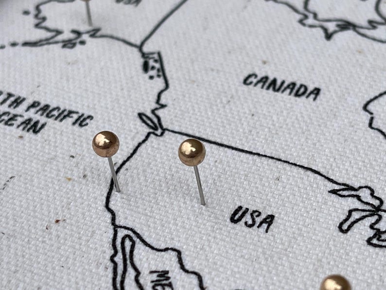 May include: A white fabric map featuring black embroidered outlines of North American countries including Canada and the USA. Two metallic gold push pins are inserted into the fabric, marking specific locations on the map surface.