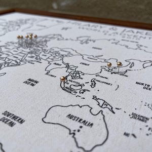 May include: A framed world map printed on white fabric with black outlines of continents and oceans. The text HILARY & JAKE'S WORLD ADVENTURES 2018 is printed in the upper right corner. Several small metallic pins are inserted into various locations.