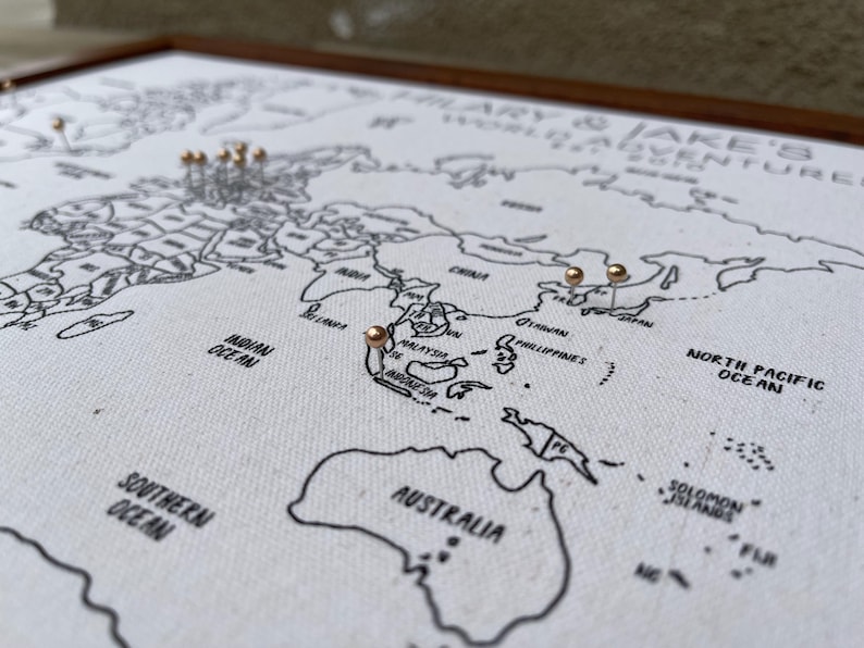 May include: A framed world map printed on white fabric with black outlines of continents and oceans. The text HILARY & JAKE'S WORLD ADVENTURES 2018 is printed in the upper right corner. Several small metallic pins are inserted into various locations.