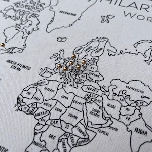 May include: A white fabric world map with black printed outlines of continents and countries. The map features gold-colored push pins inserted into various locations across Europe and North America. The text HILARY & WORLD EST is visible in the top right corner.