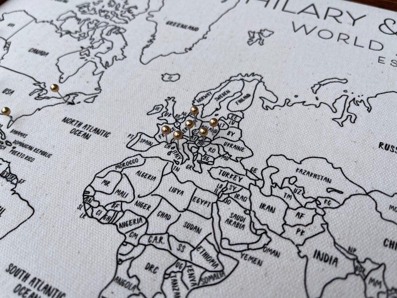 May include: A white fabric world map with black printed outlines of continents and countries. The map features gold-colored push pins inserted into various locations across Europe and North America. The text HILARY & WORLD EST is visible in the top right corner.