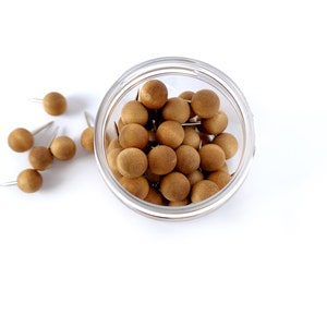 May include: A clear glass jar filled with brown wooden push pins. The push pins are scattered around the jar on a white surface.