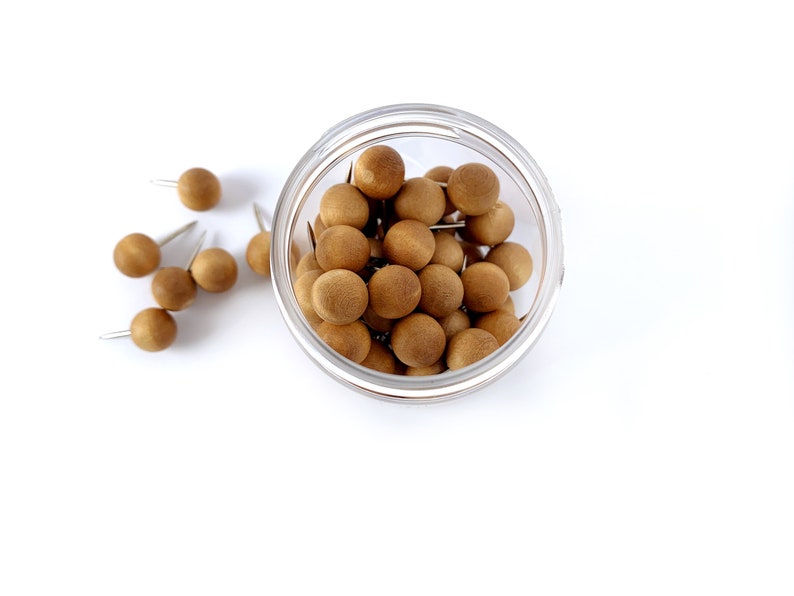 May include: A clear glass jar filled with brown wooden push pins. The push pins are scattered around the jar on a white surface.