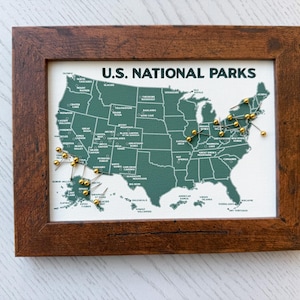 Pu&ograve; includere: Una mappa incorniciata degli Stati Uniti con la scritta "U.S. NATIONAL PARKS". La mappa &egrave; verde con scritte bianche e puntine dorate che segnano le localit&agrave;. La cornice &egrave; in legno marrone scuro.