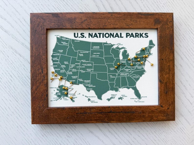 Pu&ograve; includere: Una mappa incorniciata degli Stati Uniti con la scritta "U.S. NATIONAL PARKS". La mappa &egrave; verde con scritte bianche e puntine dorate che segnano le localit&agrave;. La cornice &egrave; in legno marrone scuro.