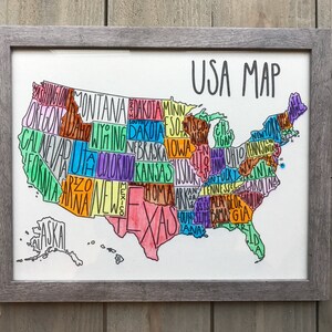 May include: A hand-drawn map of the United States of America with each state labeled in a different color. The map is framed in a gray frame and is hanging on a wooden wall.