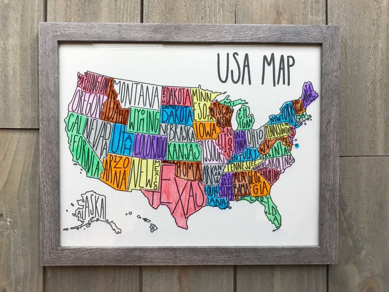 May include: A hand-drawn map of the United States of America with each state labeled in a different color. The map is framed in a gray frame and is hanging on a wooden wall.