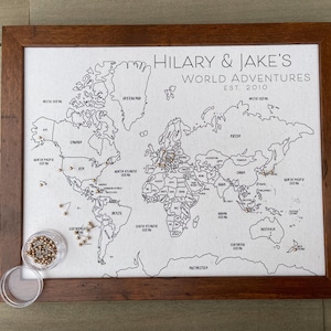 May include: A framed world map on a light fabric background with black outlines of continents and oceans. The text reads HILARY & JAKE'S WORLD ADVENTURES EST. 2010. Several metallic pins are inserted into the map, with a small clear container of extra pins.