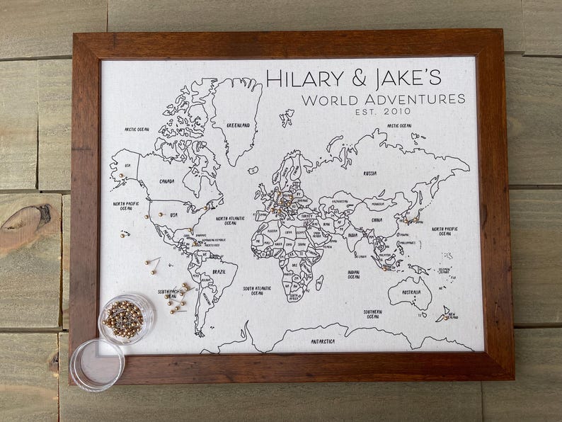 May include: A framed world map on a light fabric background with black outlines of continents and oceans. The text reads HILARY & JAKE'S WORLD ADVENTURES EST. 2010. Several metallic pins are inserted into the map, with a small clear container of extra pins.