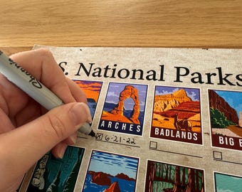 May include: A person is marking a box on a U.S. National Parks poster with a black marker. The poster features colorful illustrations of national park landmarks, including Arches and Badlands. The date 6-21-22 is written next to the checked box.