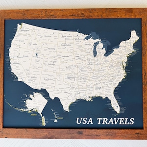 May include: Framed map of the United States with a dark blue background and a detailed white map. The map is labeled with state names and has a wooden frame. The text "USA TRAVELS" is displayed in white at the bottom.